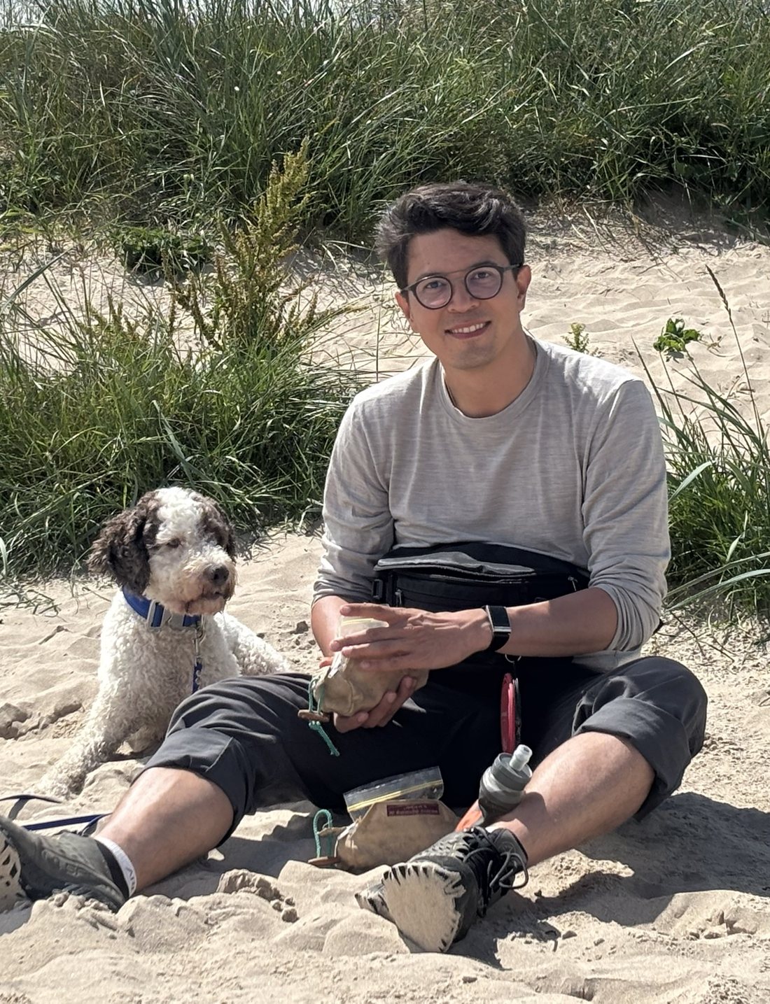 Stefan sitzt in einer Düne, den Futterbeutel in der Hand, sein Lagotto liegt neben ihm